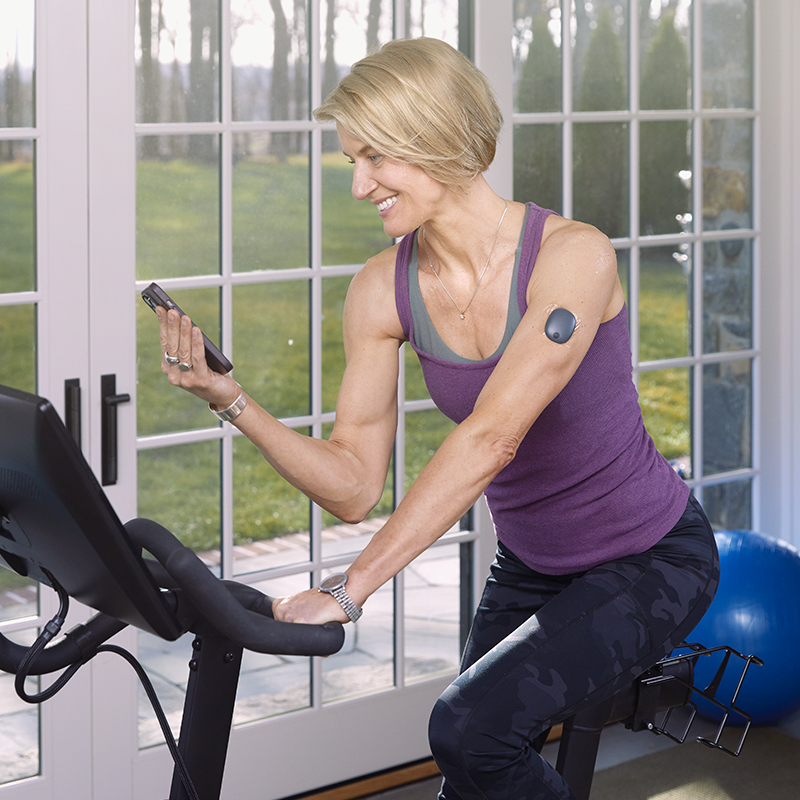 Vrouw zit op een hometrainer terwijl ze een app voor glucosemonitoring gebruikt.
