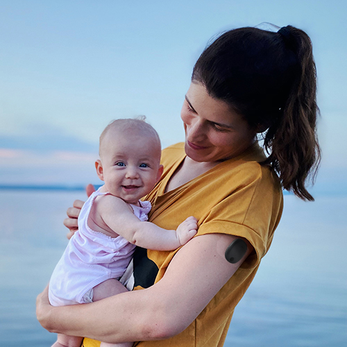 Vrouw met een Eversense langetermijn-CGM houdt haar baby vast op het strand.